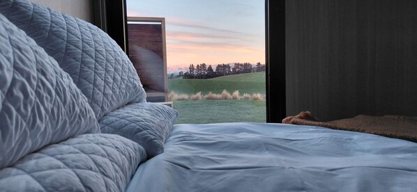 Un dormitorio con una cama grande, vistas a un campo cubierto de hierba y una estructura de madera en el exterior.