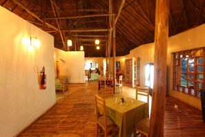 A spacious dining area with wooden floors and tables, a fire extinguisher on the wall, and a view through a window.