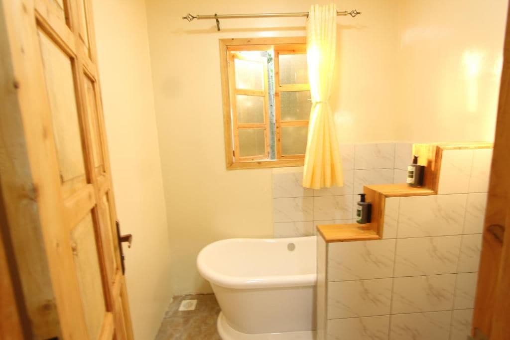 Un baño con una bañera blanca, una puerta de madera, un ventanal con cortina y un estante con artículos de aseo.