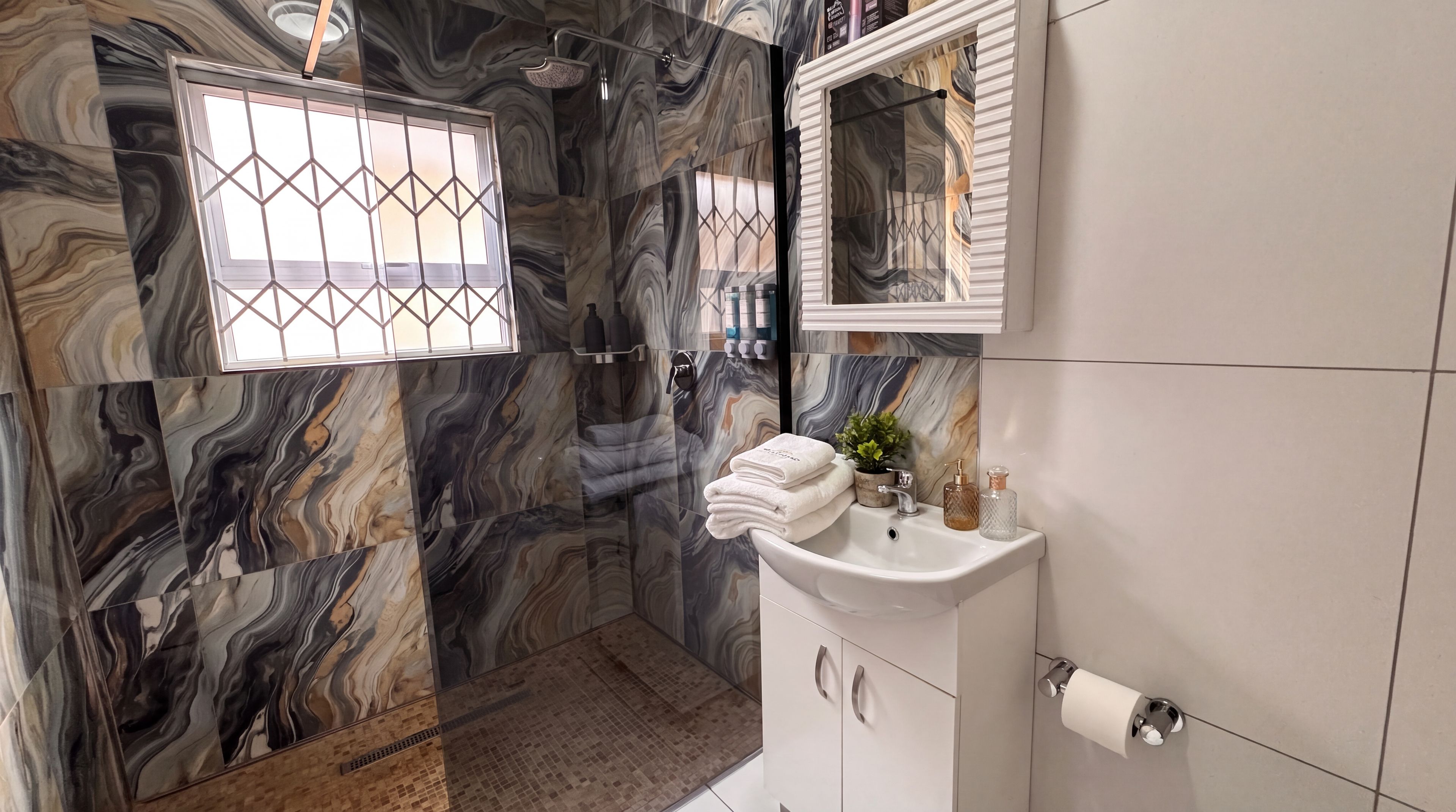 Une salle de bain avec un mur en marbre, une douche en verre, un lavabo avec des articles de toilette et un miroir.