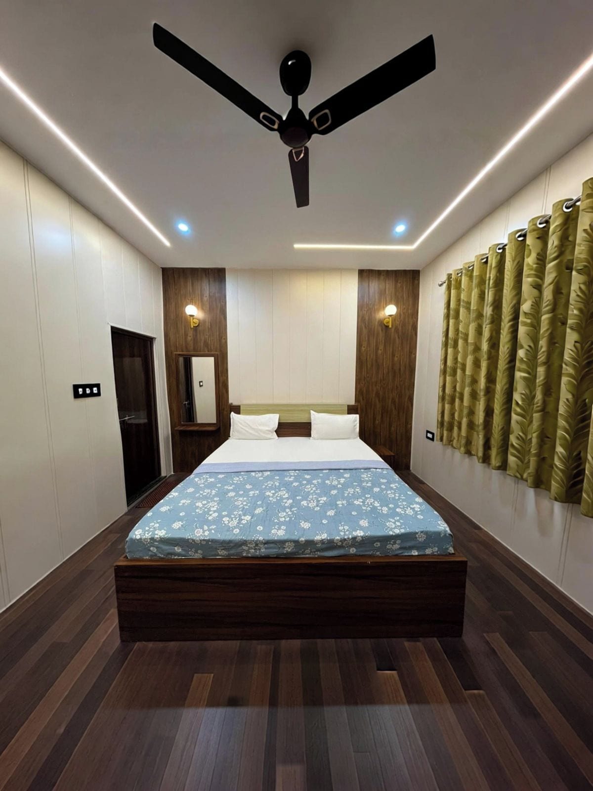 Un dormitorio con cama, ventilador de techo, iluminación empotrada, paneles de madera y una ventana con cortinas.
