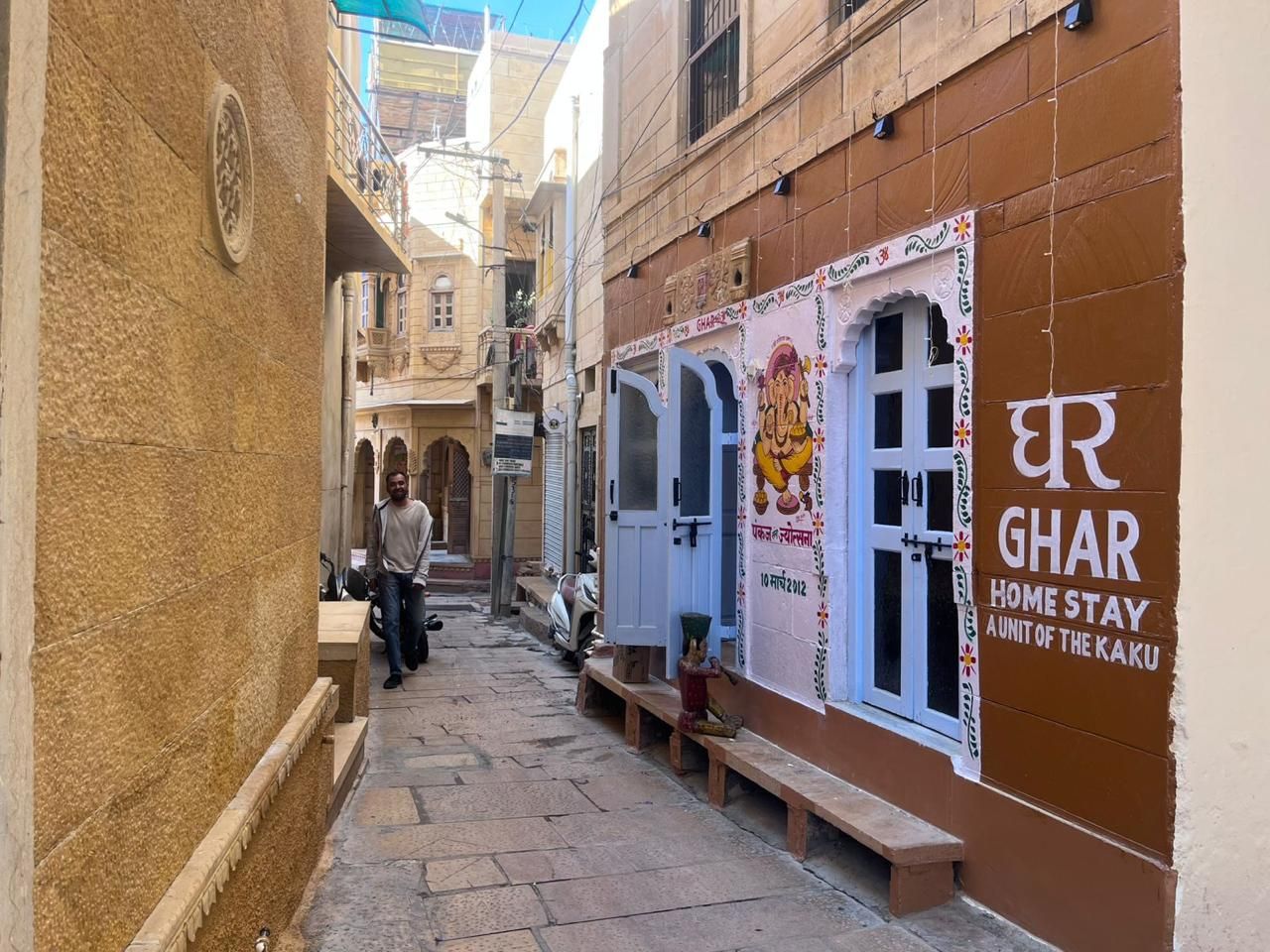 A narrow alleyway with a building on the right featuring a sign for 'Ghar Home Stay Aunit of the Kaku'.