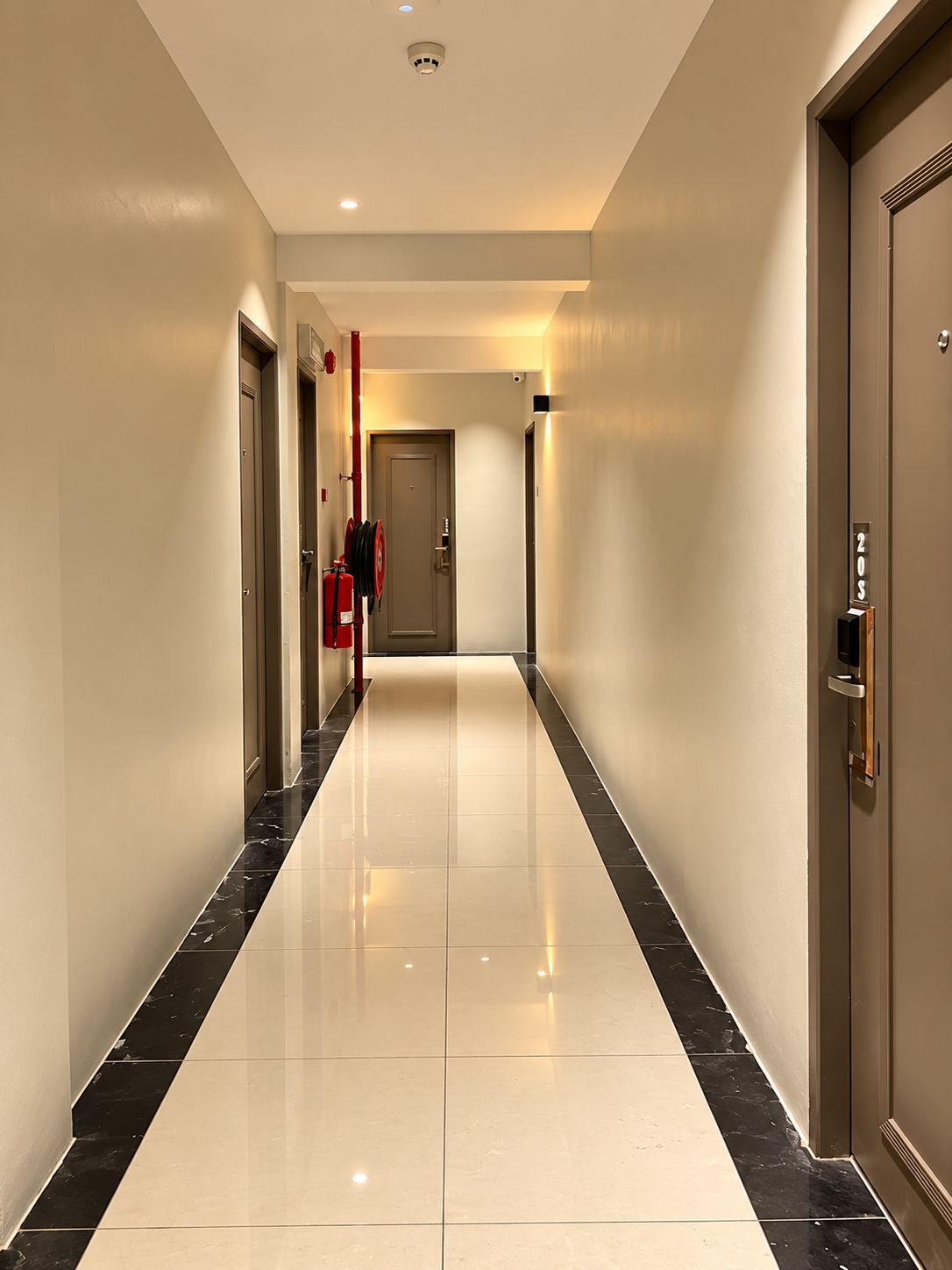 A hotel corridor with a shiny tiled floor, doors on either side, and a fire extinguisher mounted on the wall.