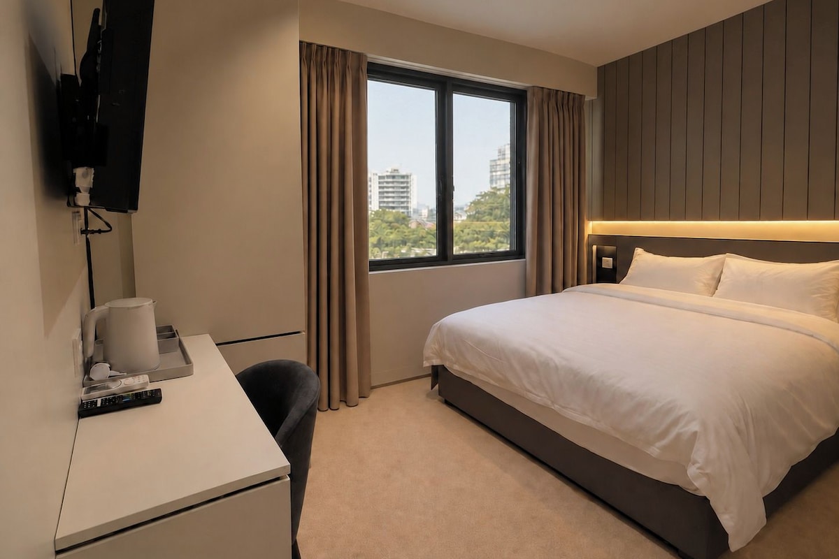 Una habitació d'hotel amb un llit, escriptori, cadira, televisió penjada a la paret i finestra amb cortines.