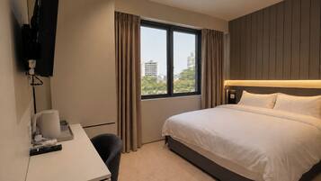 A hotel room with a bed, a desk, a chair, a television mounted on the wall, and a window with curtains.