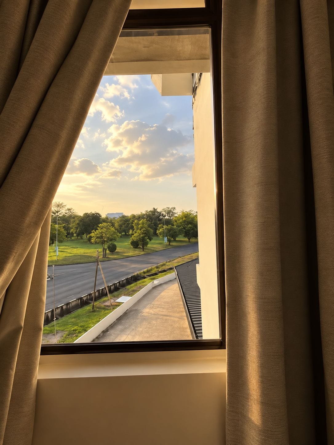 A window with curtains, a view of a road, greenery, and a clear sky.