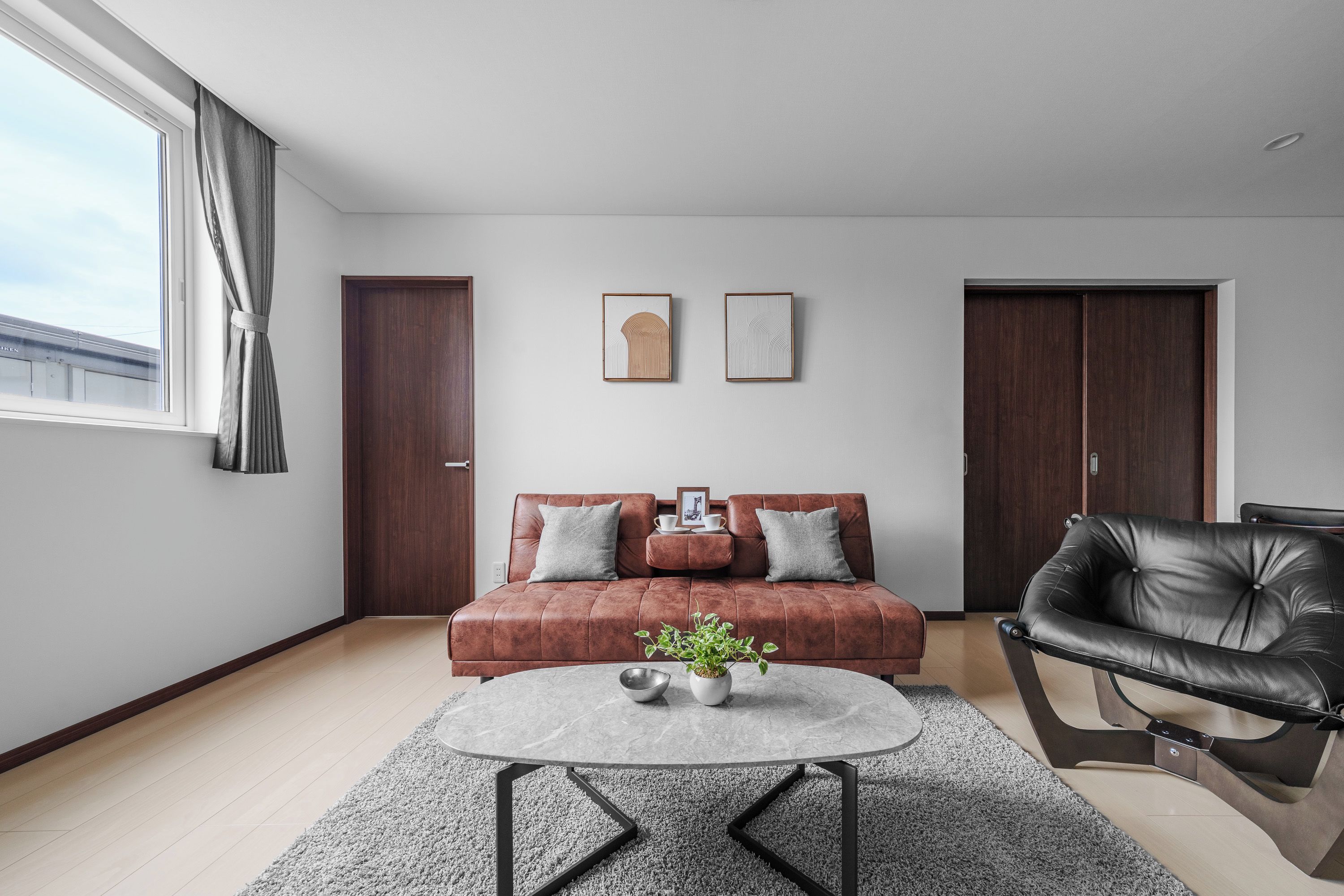 A modern living room with a brown sofa, a black leather chair, a round coffee table, and two framed artworks on the wall.