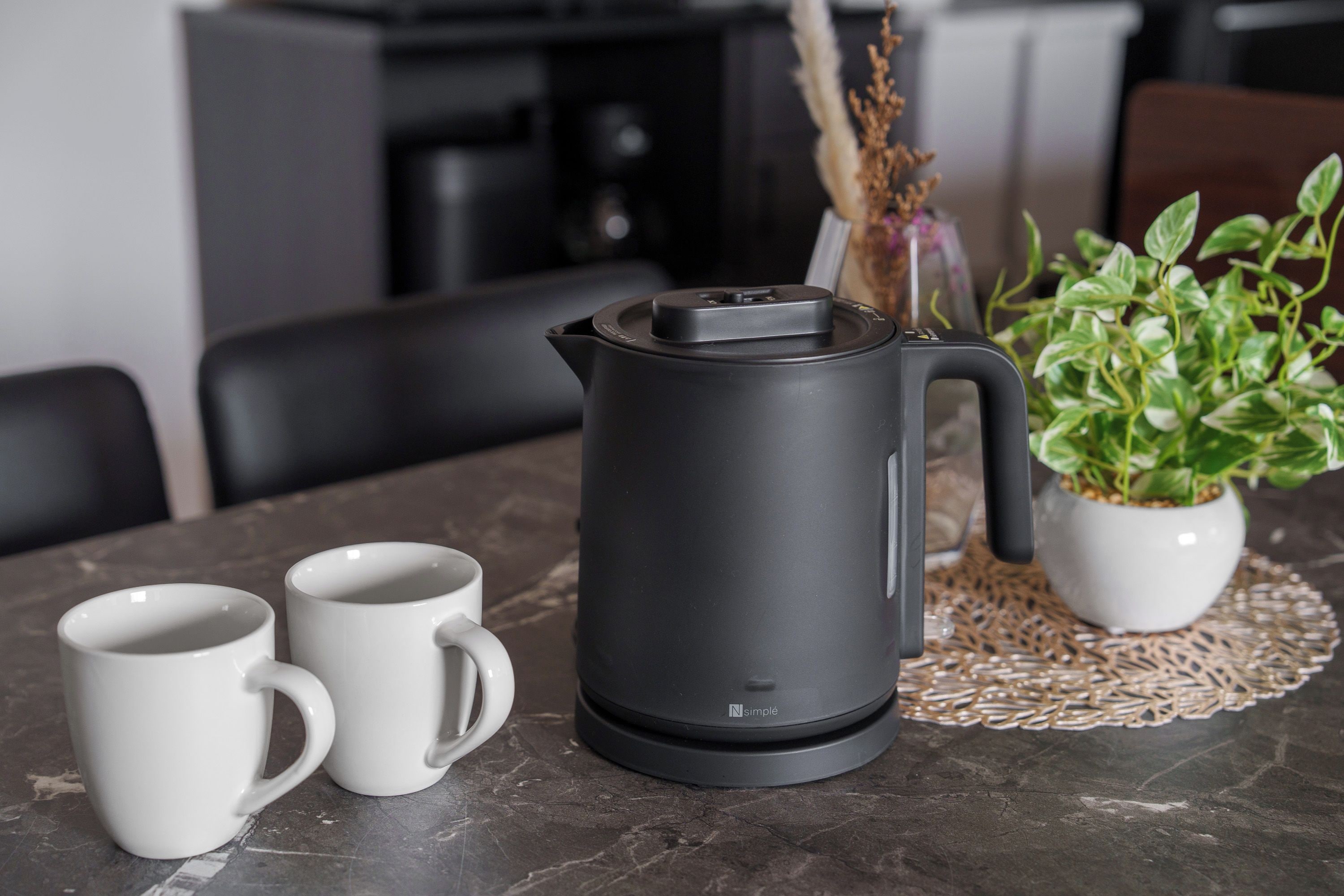A black electric kettle on a table with two white mugs beside it.