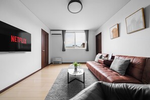 Uma sala de estar moderna com um sofá de couro, uma mesa de centro com uma planta, uma TV de tela plana exibindo 'NETFLIX' e uma janela com vista para prédios.