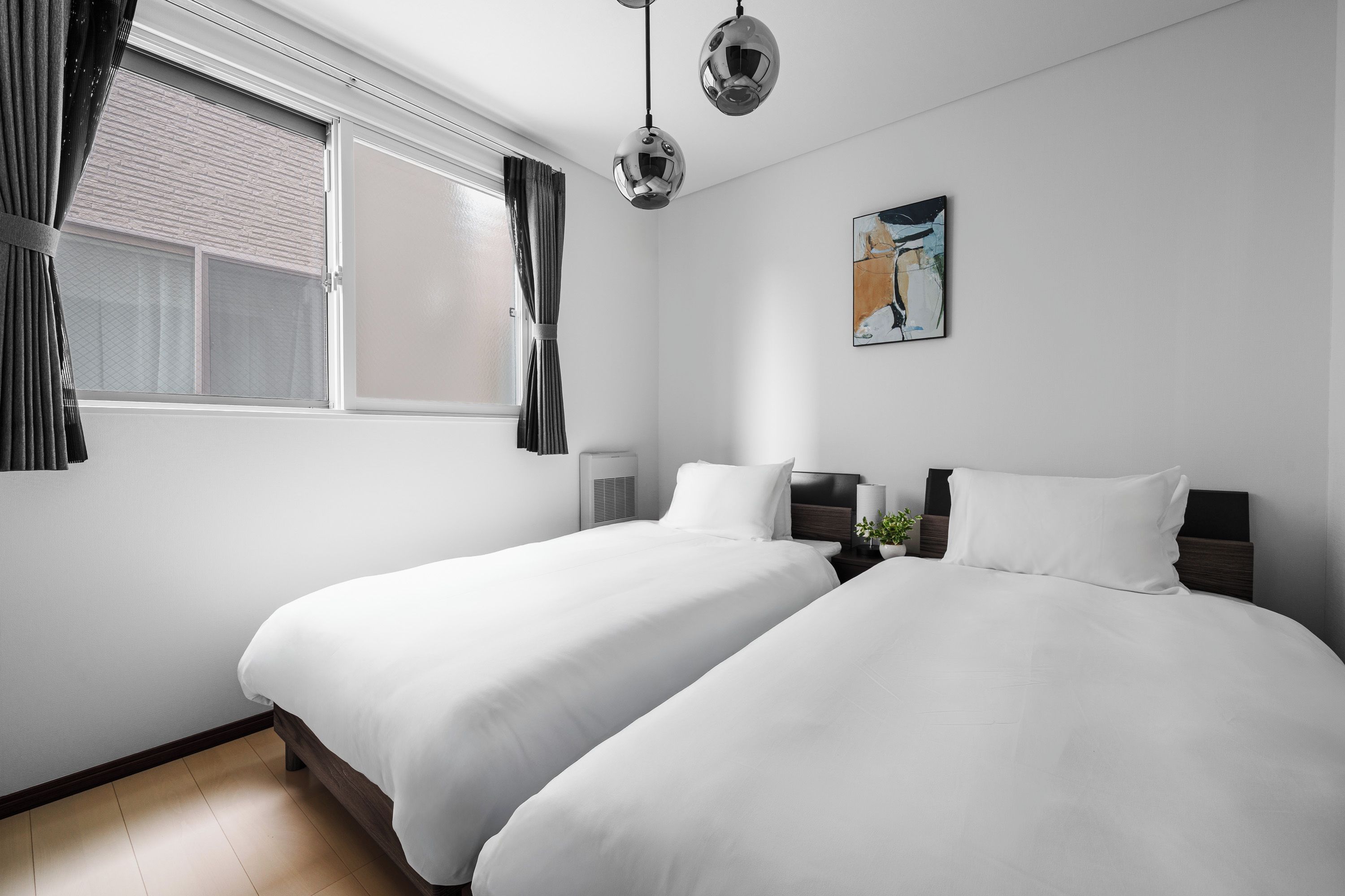 A hotel room with two beds, a window with curtains, a wall-mounted air conditioner, a painting, and a pendant light.