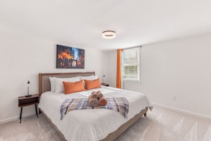 A neatly arranged bedroom with a bed, pillows, a bedside table, a lamp, a window with curtains, and a painting on the wall. - Movie Theatre | SouthPark | Patio | WFH Setup (Charlotte)