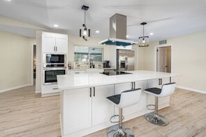 A modern kitchen with white cabinetry, a central island with bar stools, stainless steel appliances, and pendant lighting. - Beautiful Palm Coast Pool House (Palm Coast)