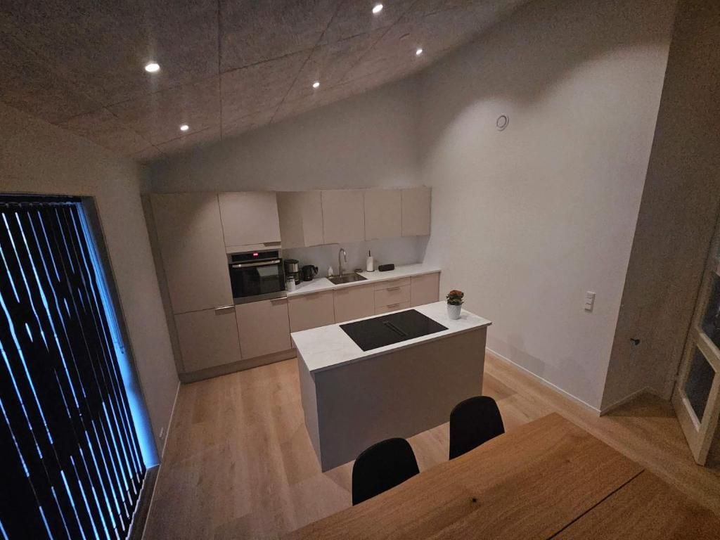Et kompakt køkken med en central ø, et spiseområde med bord og stole, samt et vindue med persienner.