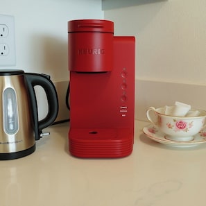 Een rode Keurig koffiezetapparaat, een roestvrijstalen Hamilton Beach koffiekan, en een bloemrijk theekopje en schoteltje op een aanrecht.
