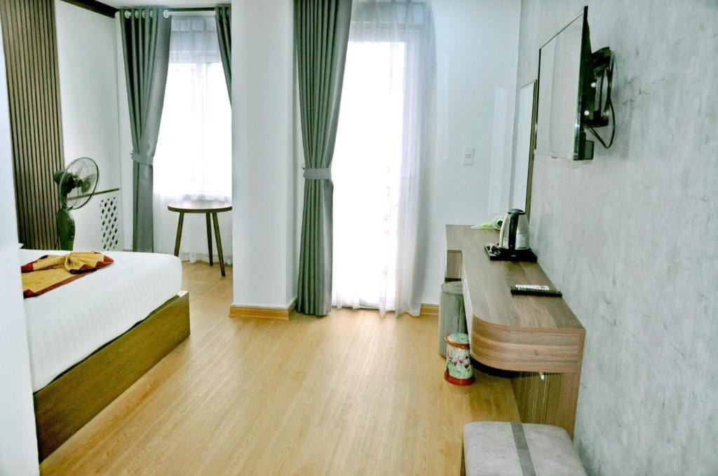 Hotelkamer met een bed, een bureau, een stoel, een tafeltje, een ventilator, een televisie en een raam met gordijnen.