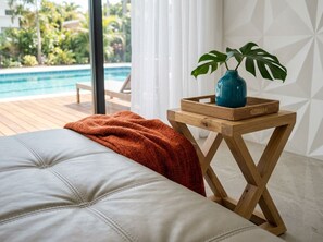Une chambre avec un grand lit, une table de chevet en bois ornée d’une plante, et une vue sur une piscine.
