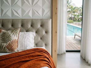Une chambre à coucher avec une tête de lit capitonnée, un lit avec un oreiller blanc et une couverture orange, et une vue sur une piscine à travers une porte vitrée.