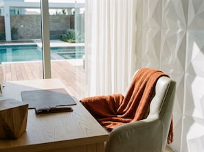 A modern room with a wooden table, a leather chair with a blanket, and a view of a swimming pool through large windows. - Spacious 4-bedroom house in tranquil Minneapolis with AC and gym (Minneapolis)