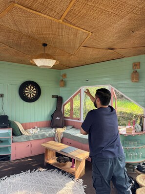 Una habitación con una pared verde, un dardo, una guitarra, una mesa de madera y una ventana con vista a vegetación.