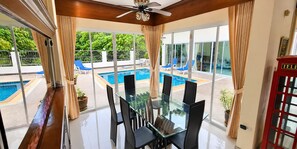 A glass dining table with chairs, a ceiling fan, and a view of a swimming pool with lounge chairs. - Private 5-bedroom Pool Villa in Middle of the Island Phuket (Kathu)