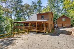 Eine Holzhütte mit Veranda, Garage und einem Schotterweg, umgeben von Bäumen.