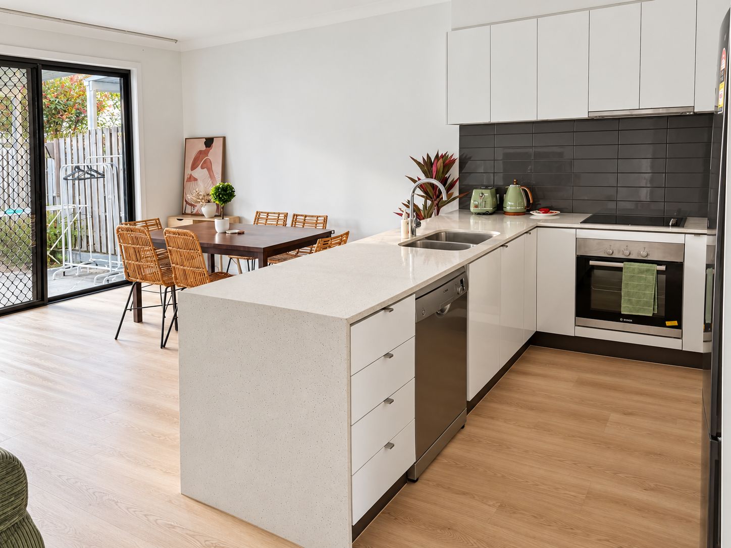 A modern kitchen with a central island, white cabinetry, a black backsplash, and a dining area with wicker chairs.