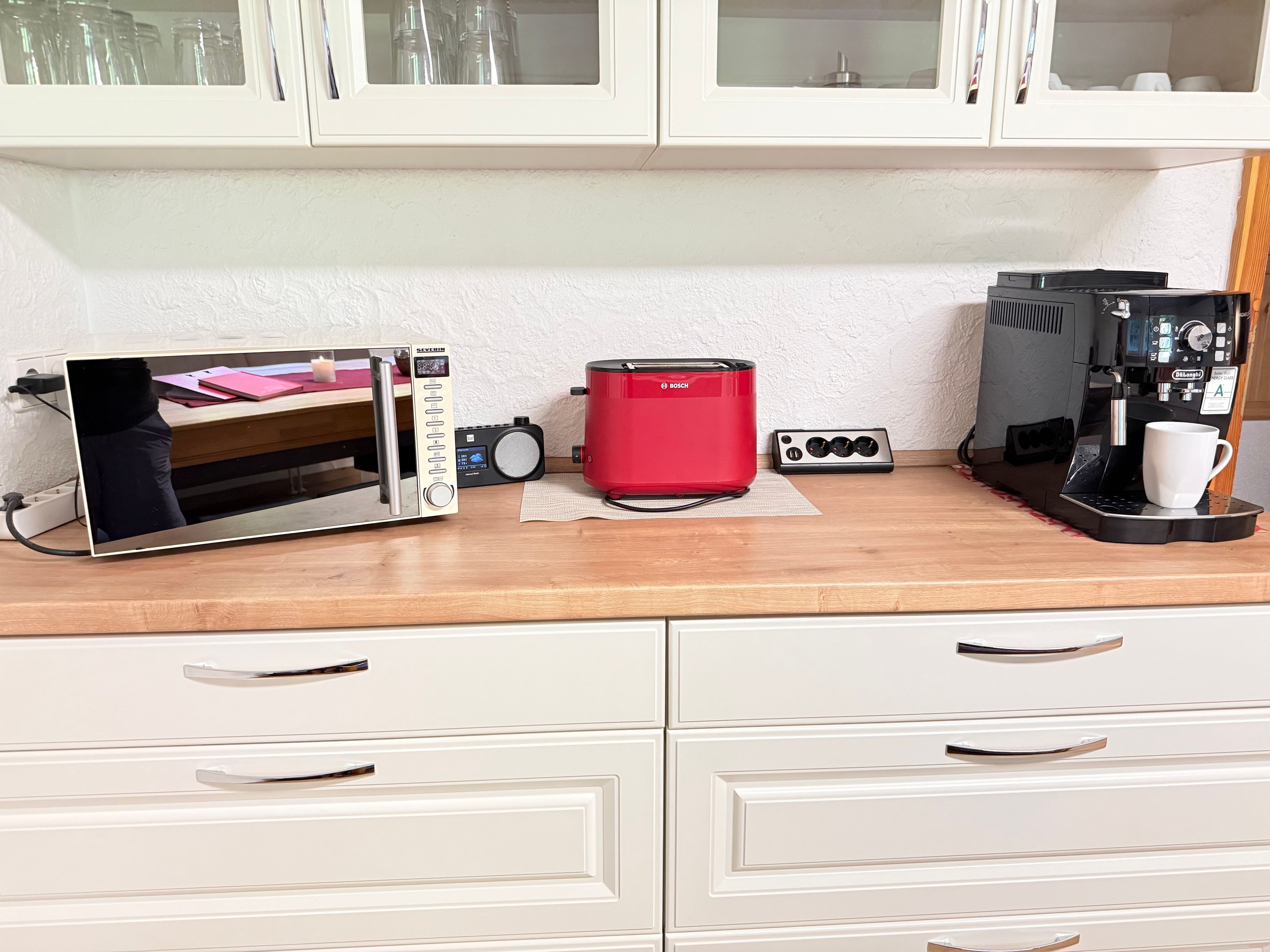 Eine Küchenzeile mit einer Mikrowelle, einem Toaster und einer Kaffeemaschine auf einer Holzausziehtafel, mit Schränken darüber.