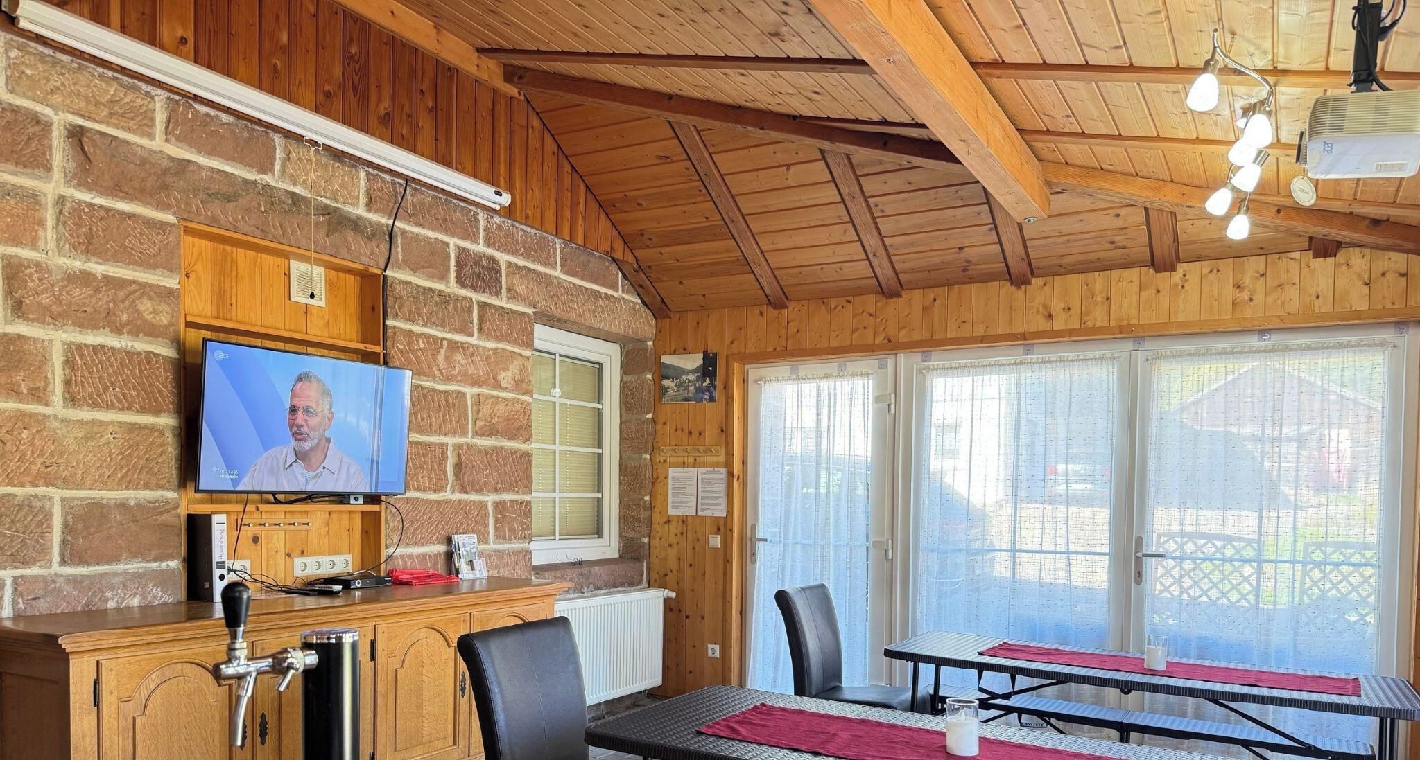 A room with a stone wall, wooden ceiling, a television mounted on the wall, a table with chairs, and a window with curtains.