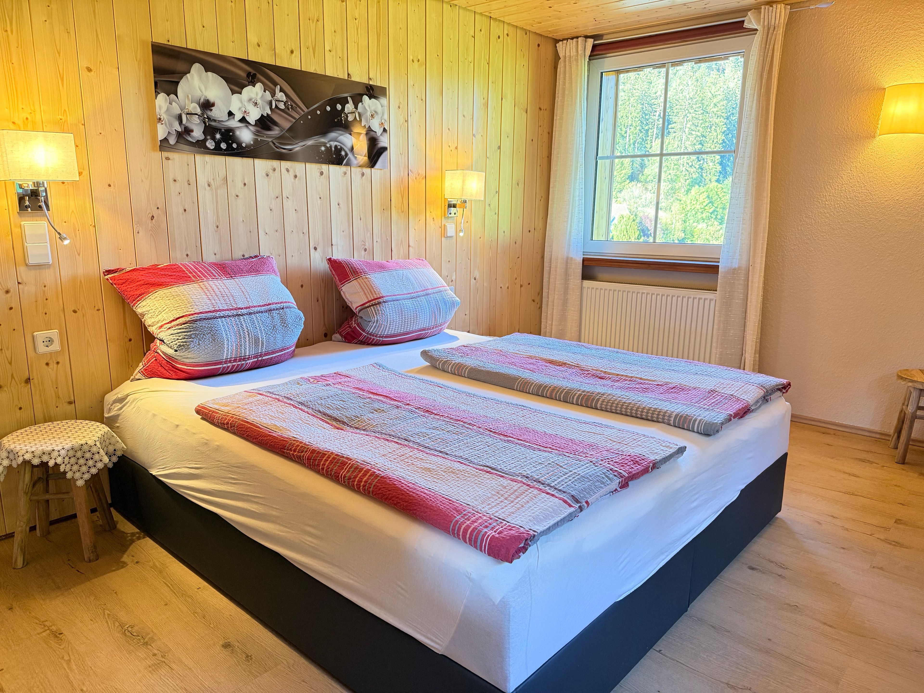 Ein Doppelbett mit gemustertem Bettbezug, zwei Kissen, ein Wandbild, Nachttischlampen und ein Fenster mit Vorhängen.
