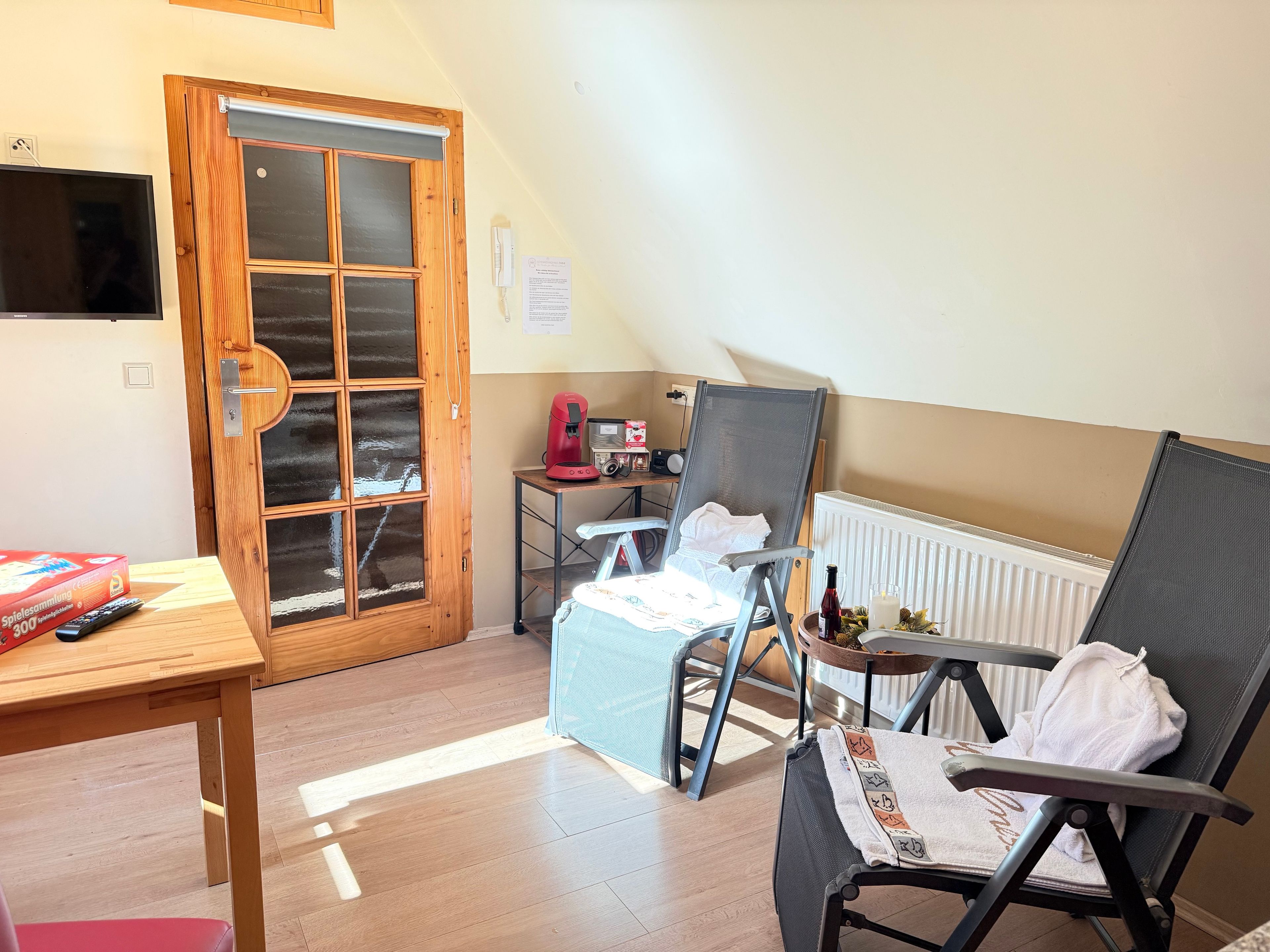 Ein Zimmer mit einer Holztür, einem Tisch mit einer roten Schachtel, einem Stuhl mit einem Handtuch, einem Heizkörper und einem kleinen Tisch mit einer Kaffeemaschine.