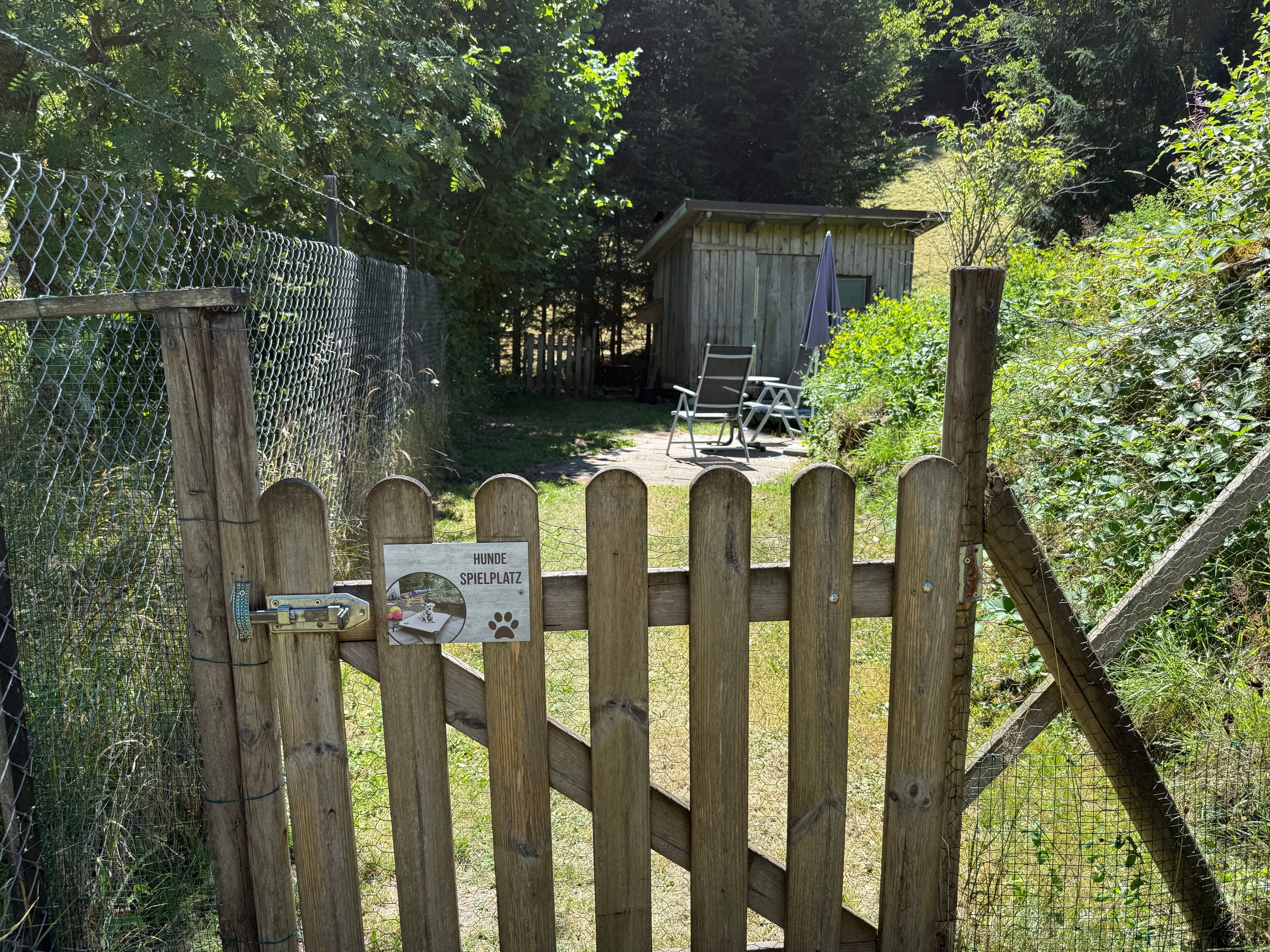 A wooden gate with a sign indicating a dog play area, a fenced enclosure, and a wooden building in the background.