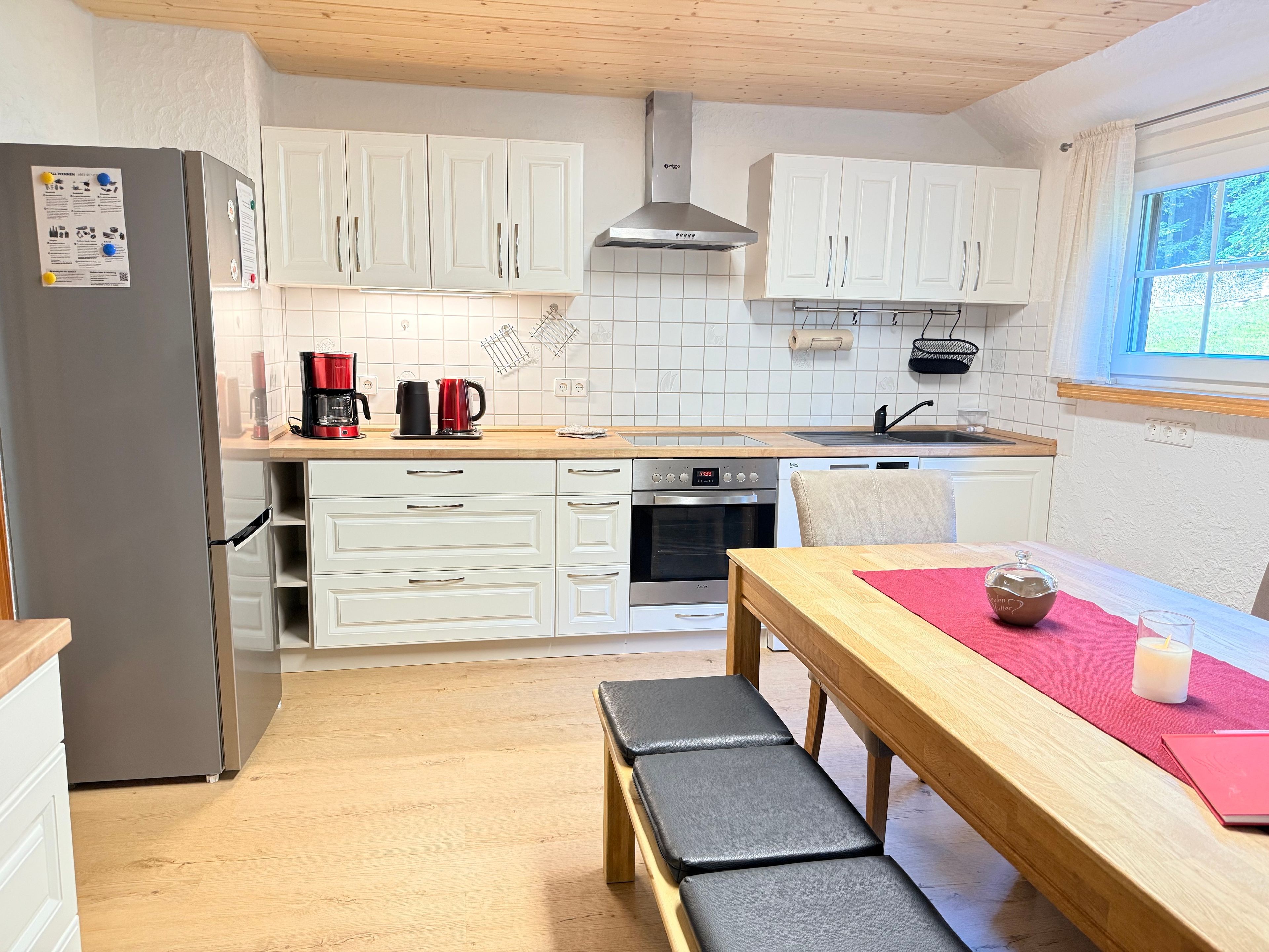 A modern kitchen with white cabinets, a stainless steel refrigerator, a coffee maker, and a dining table with a bench.