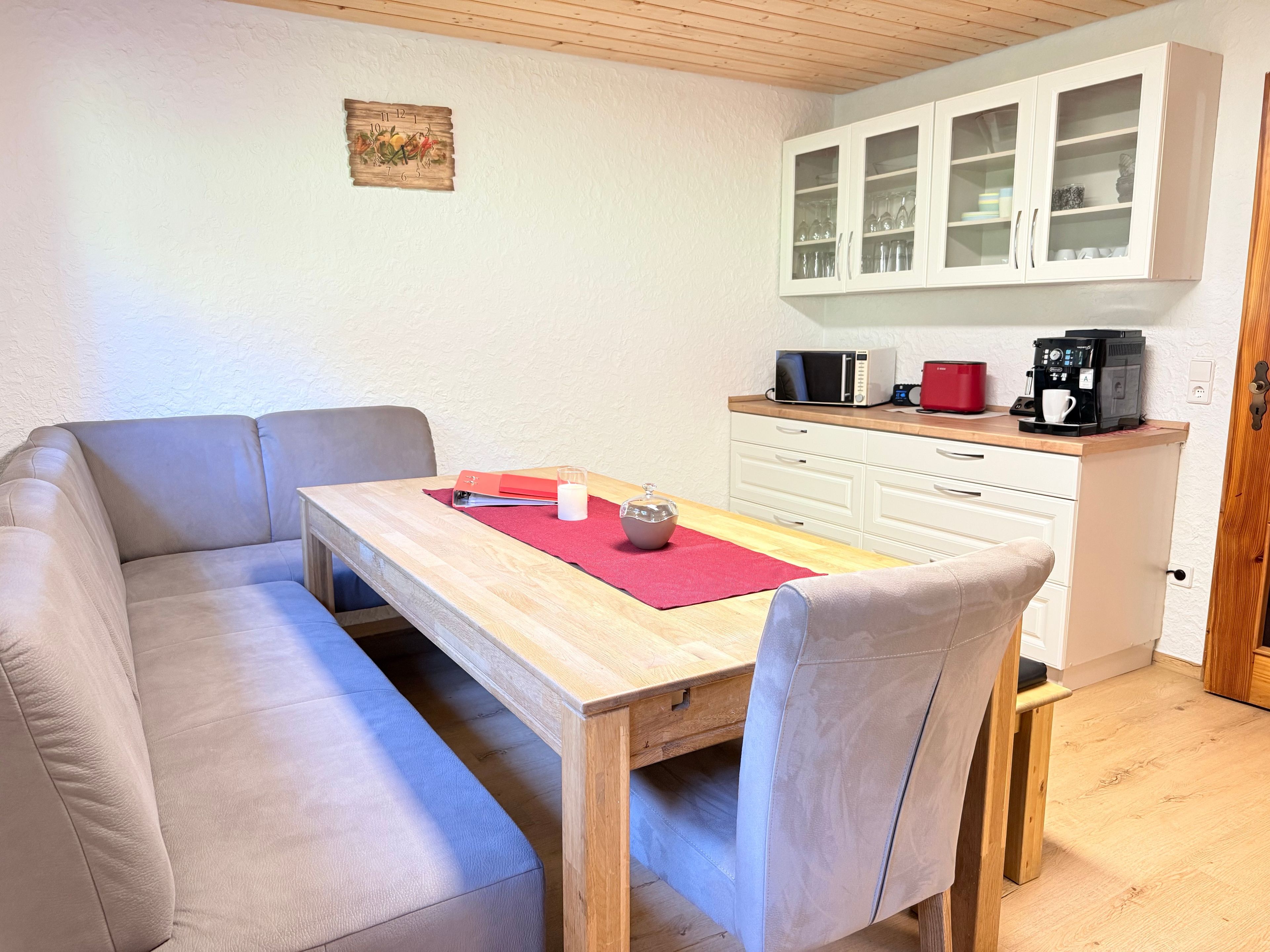 En matplats med ett träbord, två vadderade bänkar, en röd bordsduk, ett ljus och en kaffemaskin.