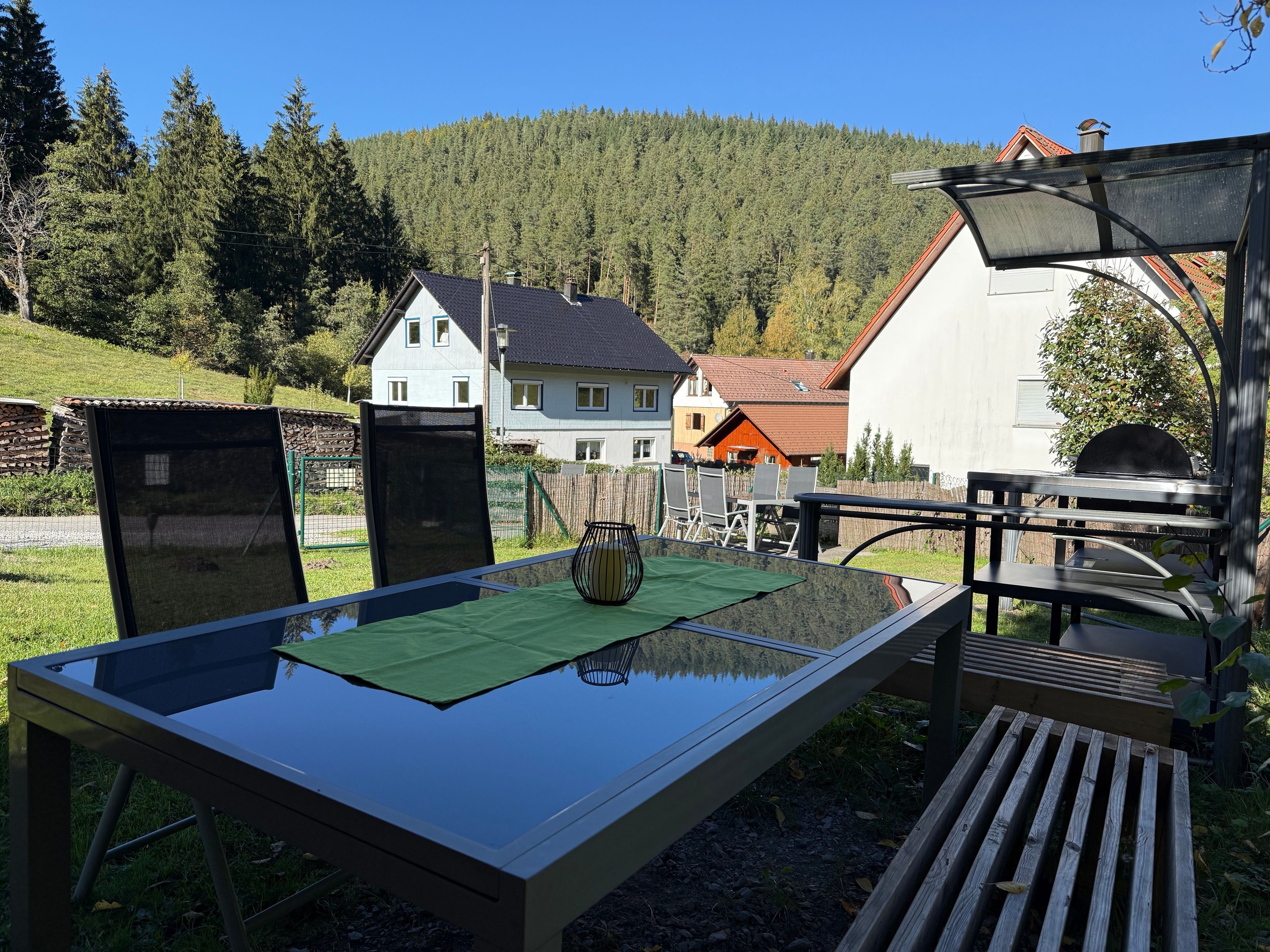 Ein Essbereich im Freien mit Tisch und Stühlen, einem Grill und Blick auf Häuser sowie einen Wald im Hintergrund.