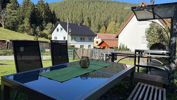 Ein Essbereich im Freien mit Tisch und Stühlen, einem Grill und Blick auf Häuser sowie einen Wald im Hintergrund.