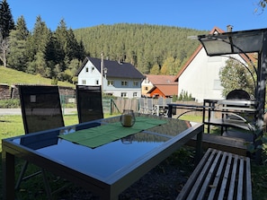 Ein Essbereich im Freien mit Tisch und Stühlen, einem Grill und Blick auf Häuser sowie einen Wald im Hintergrund.
