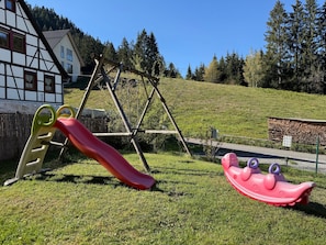 Ein Spielplatz mit Rutsche und Schaukeln, umgeben von einer Wiese und Bäumen.