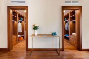 Una habitación con piso de madera, una mesa central con un florero y dos objetos decorativos, y dos armarios de madera con ropa colgada.