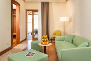 Habitación de hotel con un sofá verde, una pequeña mesa redonda con fruta, un sillón amarillo y una lámpara de pie.