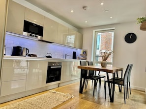 A modern kitchen with built-in appliances, a dining area with a table and chairs, and a window with a view of a brick building. - City Base: 2 Beds, 2 Baths, Balcony, Lift (Greater London)