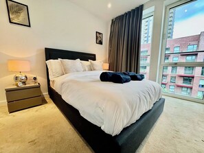 A hotel room with a large bed, a nightstand, a lamp, and a view of buildings through the window. - City Base: 2 Beds, 2 Baths, Balcony, Lift (Greater London)