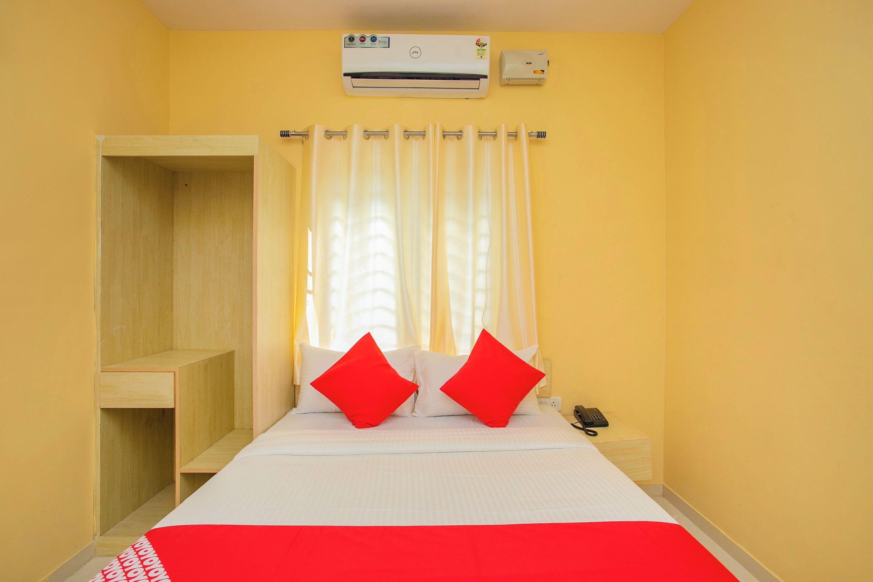 Een hotelkamer met een bed, twee rode kussens, een telefoon op een nachtkastje, een airconditioning en een raam met gordijnen.
