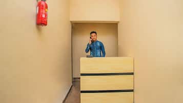 Een persoon staat achter een houten bureau in een smalle gang met beige wanden, een brandblusser aan de muur en een deur aan het einde van de gang.