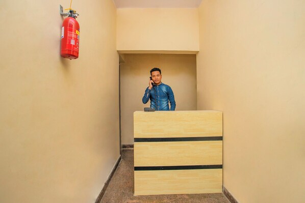 A person standing behind a wooden desk in a narrow hallway with beige walls, a fire extinguisher mounted on the wall, and a door at the end of the hallway. - Hotel O Varcity Emerald Near Ragigudda (Bangalore)
