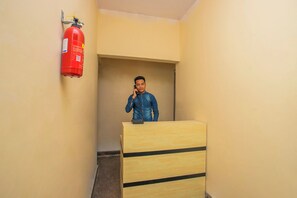 A person standing behind a wooden desk in a hallway with a fire extinguisher mounted on the wall. - Hotel O Varcity Emerald Near Ragigudda (Bangalore)