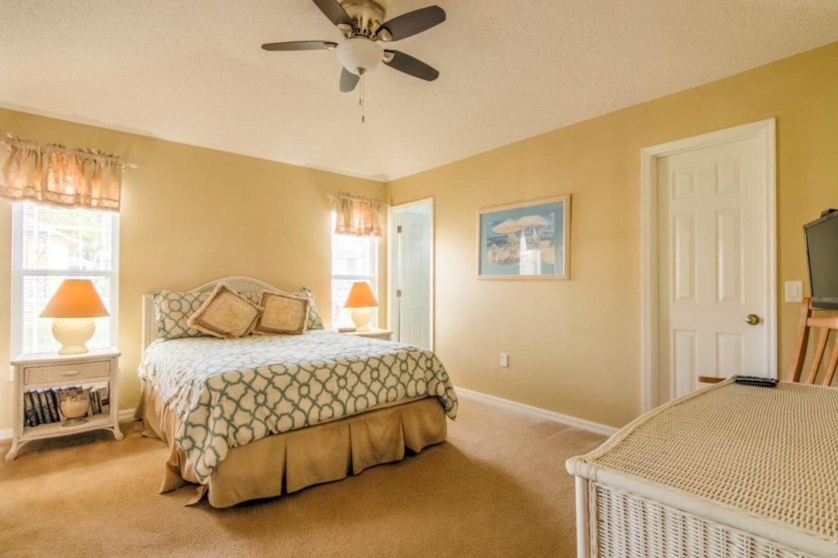 A bedroom with a bed, two bedside lamps, a ceiling fan, a door, and a wicker chair.