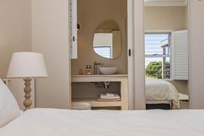 Ein Schlafzimmer mit Bett, Nachttischlampe, einem Badezimmer mit Waschbecken, Spiegel und Fenster.