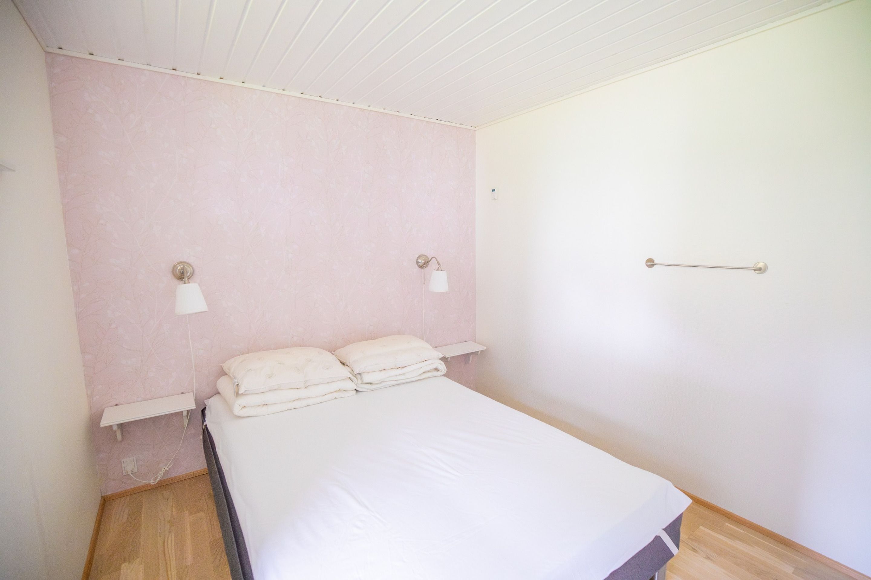 Un lit simple avec un matelas et des oreillers blancs, une table de chevet, un luminaire fixé au mur et une porte avec une poignée.