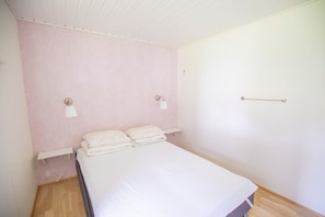 A single bed with a white mattress and pillows, a bedside table, a wall-mounted light fixture, and a door with a handle. - Eystan Gard 5H (Gjógv)