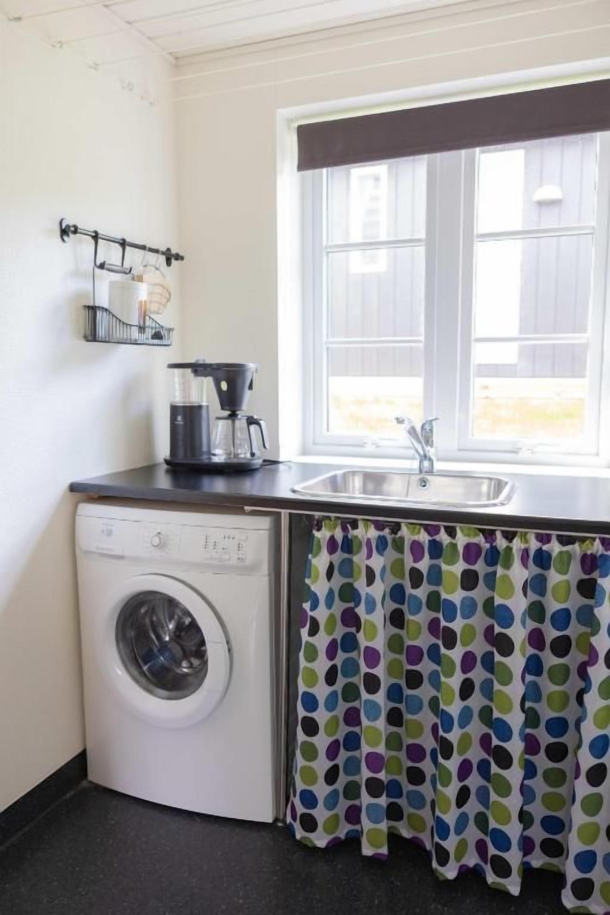 Une cuisine compacte équipée d’un lave-linge, d’une machine à café et d’une fenêtre avec des rideaux.